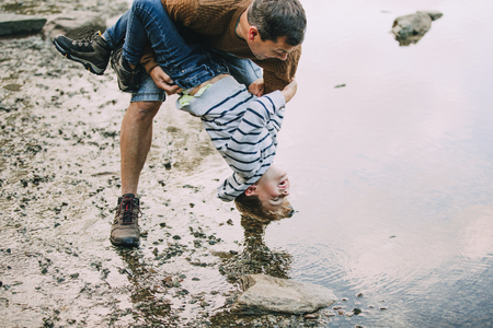 Little boy and his father have been hiking and found a pond. His father is holding the boy upside down, teasing to dunk him in the water. の写真素材