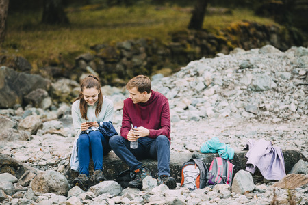 Two teenage siblings are sitting on a pebble beach after hiking. They are both looking at the girls' smart phone. の写真素材