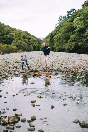 Little boy and his grandfather are out on a wlak in the woodlands. They have found a small pond and are throwing stones in for their pet dog to fetch.の写真素材