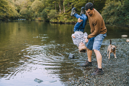 Little boy and his father have been hiking and found a pond. His father is holding the boy upside down, teasing to dunk him in the water. の写真素材