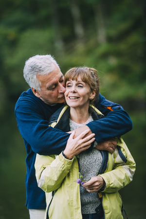 Senior couple are posing for the camera while out hiking. The man is kissing his wife with his arms wrapped round her.の写真素材