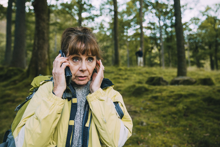 Senior couple are lost in the woods. The woman is trying to get signal on her phone and her partner can be seen in the background.の写真素材