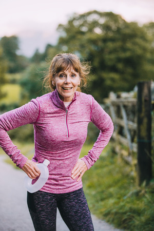 Senior woman is looking out of breath after a run through the woodland. She is standing on a track path looking at the camera with her hands on hips. の写真素材
