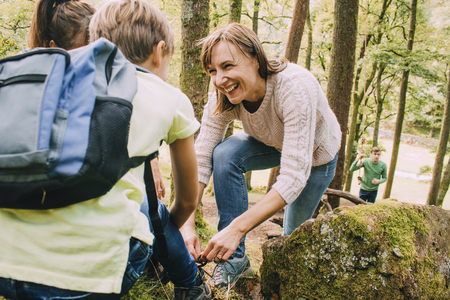 Happy family are hiking through the woodlands together. The mother has stopped to help her son tie the laces on his hiking boots.の写真素材