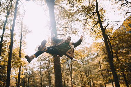 Young adult is having a fun time on a rope swing he has found while hiking in the woodlands. He has his arm and legs outstretched and is shouting in excitement. の写真素材