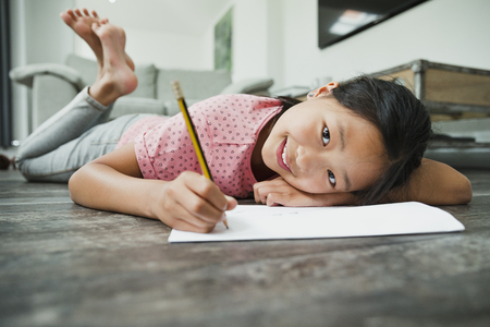 Little girl is smiling for the camera while lying on the floor doing her homework.の写真素材