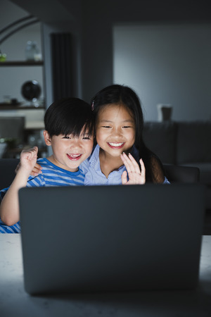 Little girl and her younger brother are using the laptop to video call their family. They are happily waving to the laptop.の写真素材
