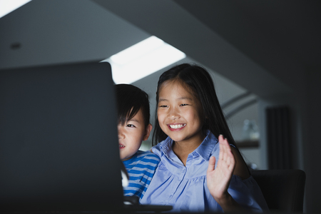 Little girl and her younger brother are using the laptop to video call their family. They are happily waving to the laptop.の写真素材