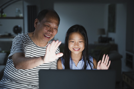 Little girl and her grandfather are using a laptop at home to video call their family. It is dark and the screen is illuminating their faces while they wave to the screen. の写真素材