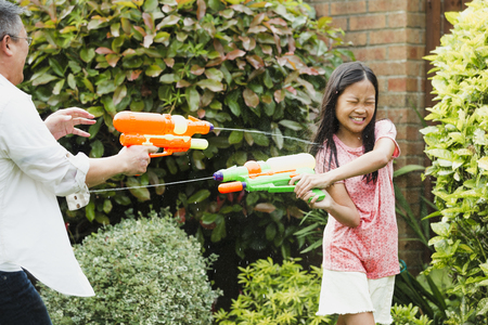 Little girl and her father are having a water fight in the garden with water pistols. The little girl is getting soaked and has her eyes closed as she tries to shoot back. の写真素材