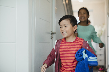 Little boy and his sister are running out the front door of their home to go swimming. The little boy has a rolled up towel in his arms.の写真素材
