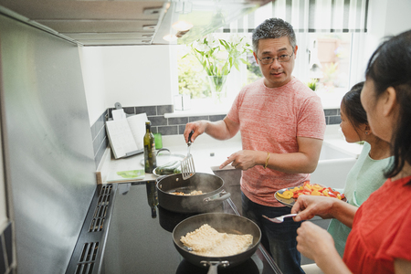 Mature man is cooking in the kitchen with his mother and daughter. He is talking to his mother about the chicken he is cooking and his daughter is standing next to him, waiting to add the vegetables she has prepared.の写真素材