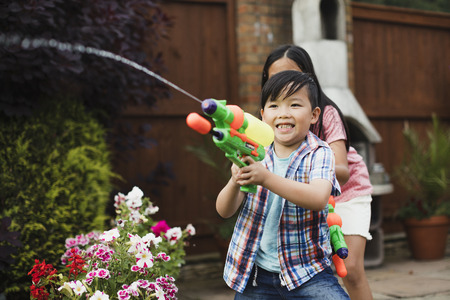 Little boy is having a water fight with his family in the garden using water pistols. His big sister is hiding behind him as he shoots.の写真素材