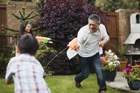 Family are having a water fight together with water pistols in the garden. The little girl is aiming for her dad, who is aiming for the little boy. の写真素材