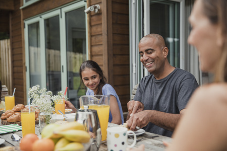 Family laughing and talking while father cuts some bread at breakfast.