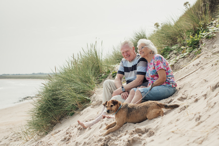 Elderly couple relaxing on the beach with their pet dog. Both sitting on a sand dune.の写真素材