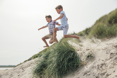 Side view of two little boys jumping over a sand dune at the beach.の写真素材