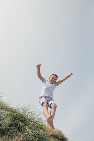 Little boy having fun and jumping over a sand dune at the beachの写真素材
