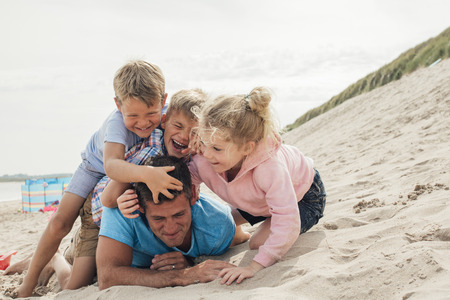 Children having fun at the beach with their father. Pining him down and tickling him.の写真素材