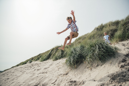 Little boy mid air, jumping over a sand dune at the beach while his brother waits at the top.の写真素材