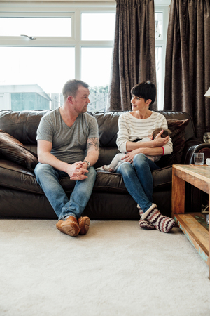 Mid adult couple are chatting on the sofa in the living room of their home while the woman is breastfeeding their baby daughter. の写真素材