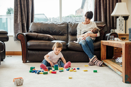 A mother is watching her daughter play with toy blocks in the living room as she breastfeeds the baby on the sofa. の写真素材