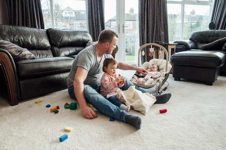 Stay at home father with his daughters at home. The eldest girl is sitting with her father playing with toy dinosaurs while the baby girl is in her rocker chair next to them.の写真素材