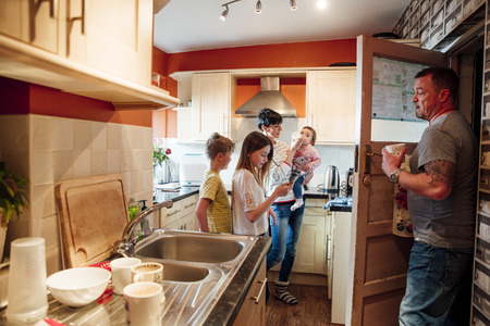 A whole family are busy in the kitchen of their home. The mother is feeding the baby while the father does the dishes. The older girl is using a smartphone and her brother is watching the baby get fed. の写真素材