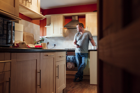 Mature man is standing in the kitchen of his home with a cup of tea in his hand, staring deeply out of the window. の写真素材