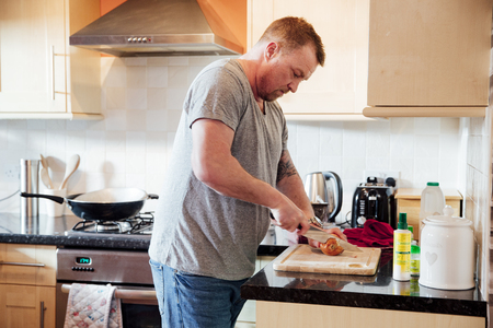Mature man is cooking in the kitchen of his home. He is cutting up an onion on a chopping board. の写真素材