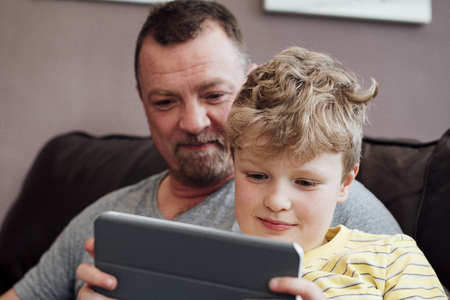 Little boy is sitting on his dads knee on the sofa at home, using a digital tablet. His dad is watching him.の写真素材