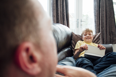 Little boy and his father are talking while lying on the sofa together. The little boy is holding a digital tablet.の写真素材