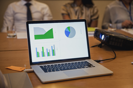 Front view of a computer plugged into a projector showing charts and graphs with information on. Unrecognisable people in the background.の写真素材