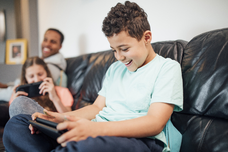 Little boy is playing on a handheld game console at home with his sister and their father is watching them.の写真素材