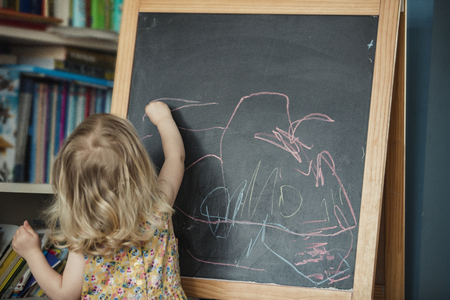 Rear view of a little girl drawing and writing on a blackboard. The little girl is reaching up and drawing, using different coloured chalk on the black board.の写真素材