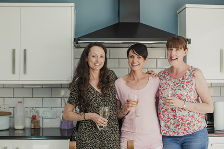 Three mid adult women standing in a kicthen enjoying a glass of wine. The women are all standing...