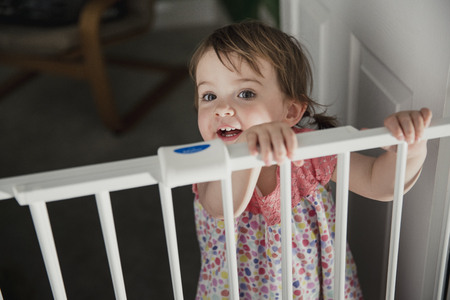 Front view of a little girl standing on the other side of a baby saftey gate. The little girl is holding onto the top of the saftey gate and looking at the camera over the top.の写真素材