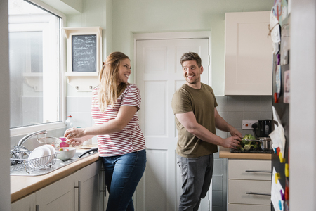 Mid-adult woman standing in the kitchen at home rinsing strawberries to clean them. Her husband is standing behind and looking at her while chopping some broccoli.の写真素材