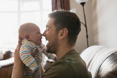 Father holding his new born baby boy up to his face to make him laugh. They are sitting in the living room on the sofa.の写真素材