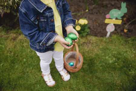 HIgh angle view of an unrecognsiable child. They are holding their hands together, cupped, with chocolate easter eggs inside. She has a basket at her feet with some more easter eggs in.の写真素材