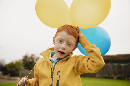 Headshot of a little boy looking at the camera while holding two yellow balloons and one blue balloon.の写真素材
