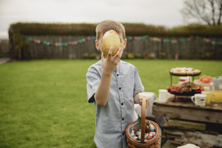 Little boy holding a chocolate easter egg infront of his face. He is standing outdoors and holding a...