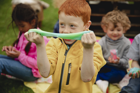 Excited little boy sitting outdoors with his friends surrounding him. He is amazed by the slime in a...