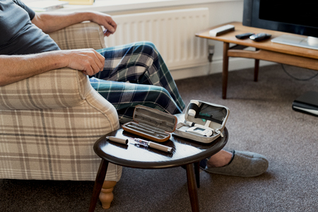 Senior man is relaxing in the living room of his home with a blood glucose testing kit on the table beside him.の写真素材