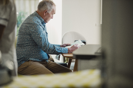 Senior diabetic man is sitting at a dining table reading a newspaper while his carer prepares his medication.の写真素材