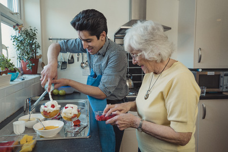 Teenage boy making fruit compote with his grandmother in the kitchen of her home. He is adding squirty cream to the bowls.の写真素材