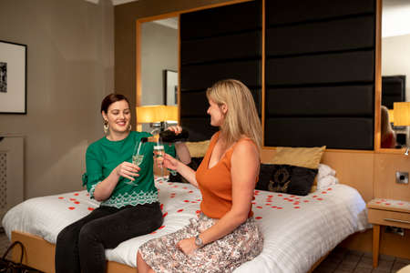 Two caucasian women drinking champagne, celebrating and having fun together on a hotel bed in Jesmond, England.の写真素材
