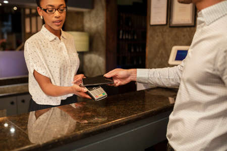 A female hotel receptionist taking the contactless payment of a customer who is checking in to the hotel, he is using his smart phone to pay.の写真素材