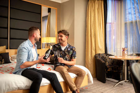 Two caucasian men drinking champagne, celebrating and having fun together on a hotel bed in Jesmond, England.の写真素材