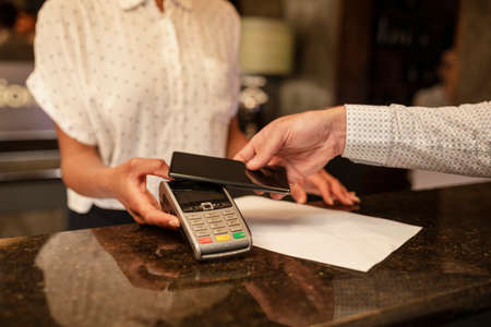 An unrecognisable female hotel receptionist taking the contactless payment of a customer who is checking in to the hotel, he is using his smart phone to pay.の写真素材